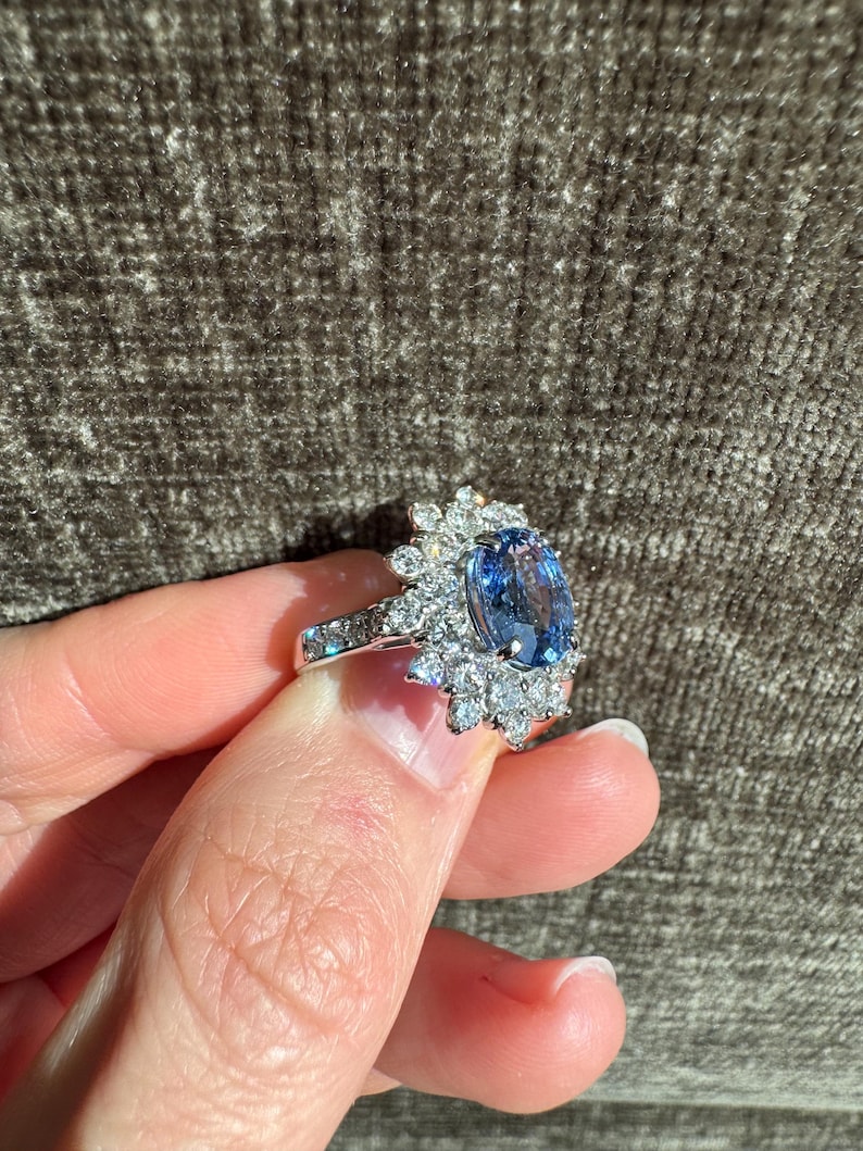 May include: A silver ring featuring a large, oval-cut blue gemstone surrounded by smaller, clear gemstones. The ring's band is also adorned with smaller, clear gemstones. The ring is held up against a textured, brown background.