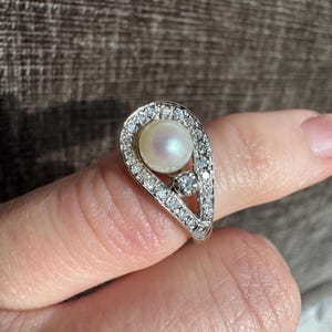 May include: A silver ring featuring a pear-shaped design. The ring is adorned with a central pearl, surrounded by a halo of small, clear gemstones. A smaller gemstone is set below the pearl.