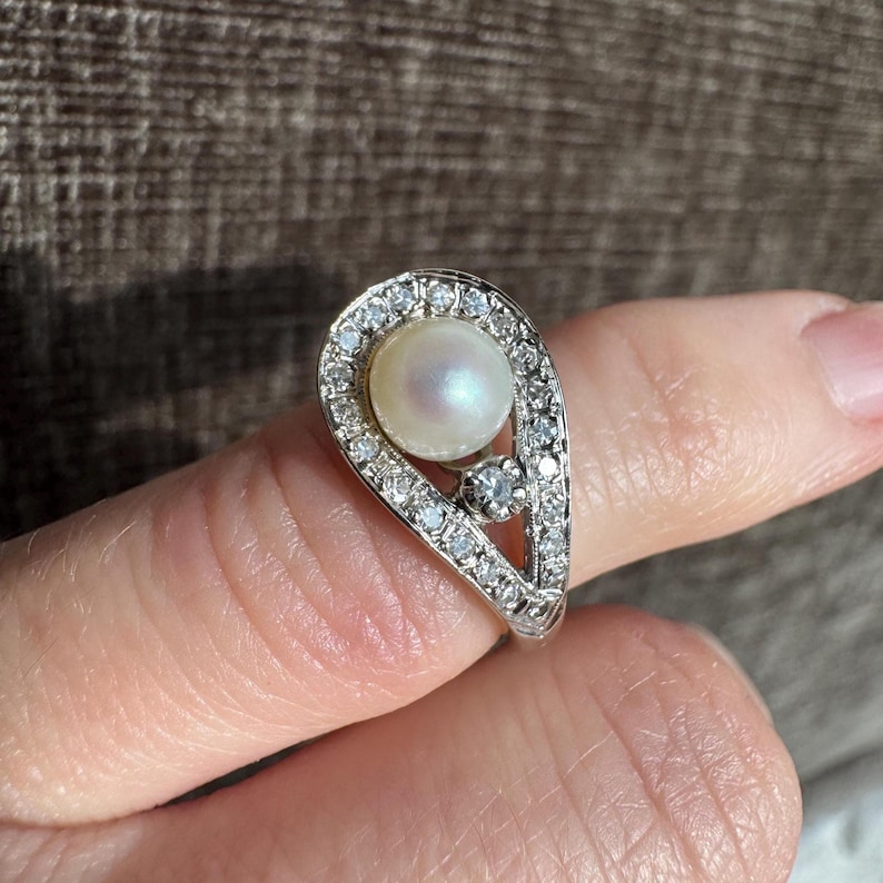 May include: A silver ring featuring a pear-shaped design. The ring is adorned with a central pearl, surrounded by a halo of small, clear gemstones. A smaller gemstone is set below the pearl.