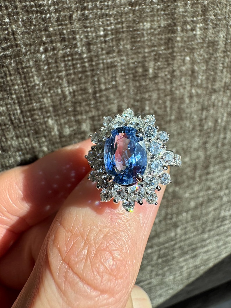 May include: A silver ring featuring a large, oval-cut blue gemstone surrounded by smaller, clear gemstones. The ring's design is a cluster of sparkling stones around the central blue gem, creating a halo effect. The ring is held up against a neutral background.