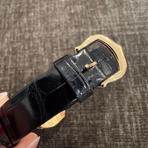 Puede incluir: Primer plano de una correa de reloj de cuero negro con una hebilla dorada. La correa tiene una superficie texturizada y la hebilla es rectangular con bordes redondeados. El reloj parece ser un art&iacute;culo de lujo.