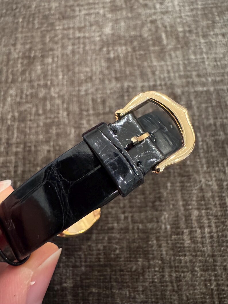 Puede incluir: Primer plano de una correa de reloj de cuero negro con una hebilla dorada. La correa tiene una superficie texturizada y la hebilla es rectangular con bordes redondeados. El reloj parece ser un art&iacute;culo de lujo.