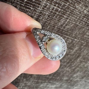 May include: A teardrop-shaped pendant featuring a central pearl surrounded by a halo of small, sparkling diamonds. The pendant is set in a silver or white gold metal, creating a classic and elegant design.