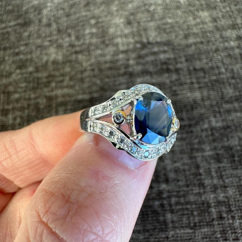 May include: A silver ring featuring a large, oval-cut, deep blue gemstone as the centerpiece. The ring is adorned with smaller, clear gemstones and gold accents, creating a luxurious and elegant design. The ring is held up by a finger.