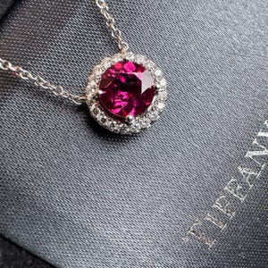 Può includere: Una collana d'argento con un pendente rotondo di pietra preziosa rosa intenso, circondato da piccole pietre preziose trasparenti. Il pendente è appeso a una delicata catena. La collana è presentata su una superficie grigio scuro.