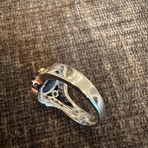 May include: A silver ring with a dark blue gemstone and small clear stones. The ring has a thick band and a detailed setting around the central stone. A small gold accent is visible.