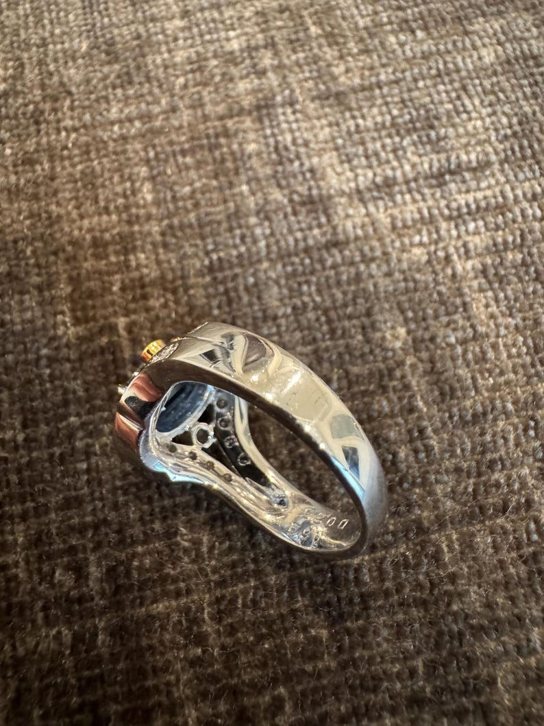 May include: A silver ring with a dark blue gemstone and small clear stones. The ring has a thick band and a detailed setting around the central stone. A small gold accent is visible.