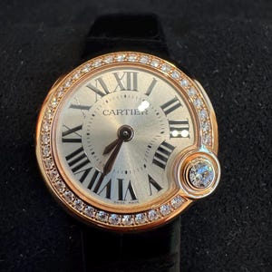 Puede incluir: Un reloj de lujo con esfera redonda dorada, n&uacute;meros romanos y la marca Cartier. El bisel est&aacute; engastado con peque&ntilde;os diamantes. El reloj tiene una correa de cuero negro y un peque&ntilde;o detalle de diamante. Hecho en Suiza.
