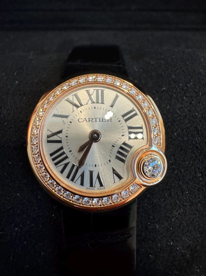 Puede incluir: Un reloj de lujo con esfera redonda dorada, n&uacute;meros romanos y la marca Cartier. El bisel est&aacute; engastado con peque&ntilde;os diamantes. El reloj tiene una correa de cuero negro y un peque&ntilde;o detalle de diamante. Hecho en Suiza.