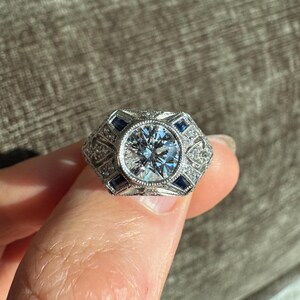 K&ouml;nnte beinhalten: Ein silberner Ring mit einem gro&szlig;en, runden, klaren Edelstein in der Mitte. Der Ring hat ein geometrisches Design mit kleinen, quadratischen blauen Edelsteinen und kleineren, klaren Steinen. Der Ring wird von einem Finger gehalten.