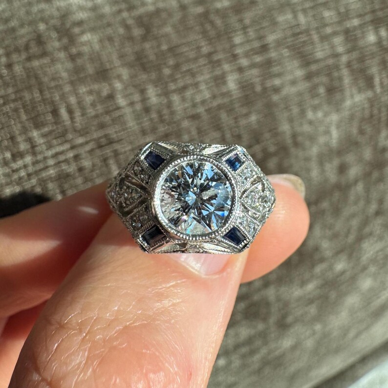 K&ouml;nnte beinhalten: Ein silberner Ring mit einem gro&szlig;en, runden, klaren Edelstein in der Mitte. Der Ring hat ein geometrisches Design mit kleinen, quadratischen blauen Edelsteinen und kleineren, klaren Steinen. Der Ring wird von einem Finger gehalten.