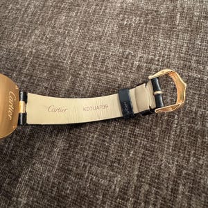 Puede incluir: Primer plano de una correa de reloj de cuero negro con una hebilla dorada. La marca "Cartier" est&aacute; impresa en la correa, junto con el n&uacute;mero de serie KD7UAP39. La correa del reloj est&aacute; sobre una superficie marr&oacute;n texturizada.