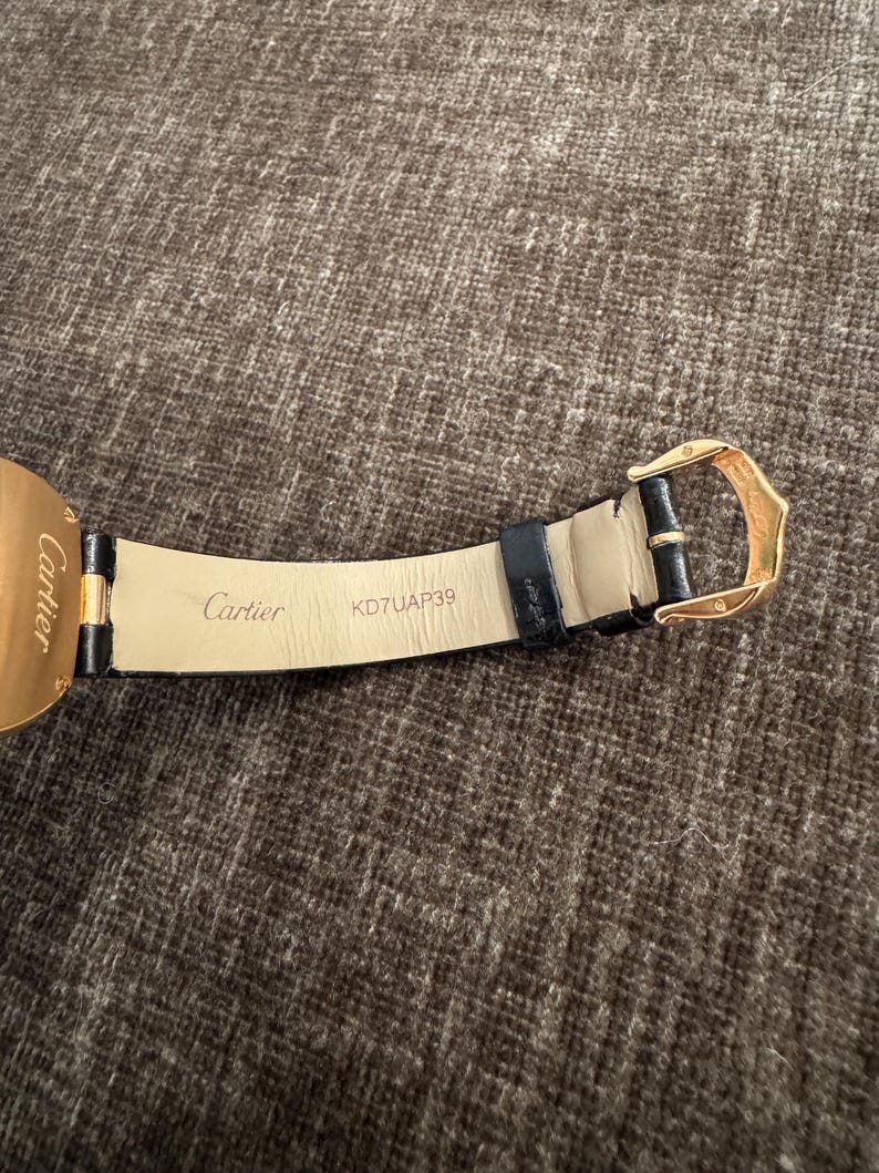 Puede incluir: Primer plano de una correa de reloj de cuero negro con una hebilla dorada. La marca "Cartier" est&aacute; impresa en la correa, junto con el n&uacute;mero de serie KD7UAP39. La correa del reloj est&aacute; sobre una superficie marr&oacute;n texturizada.