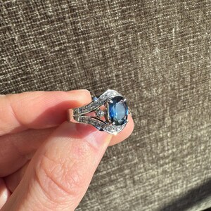 May include: A silver ring featuring a large, oval-cut, deep blue gemstone as the centerpiece. The ring is adorned with smaller, clear gemstones along the band and around the main stone, creating a sparkling effect. The ring is held up against a brown background.