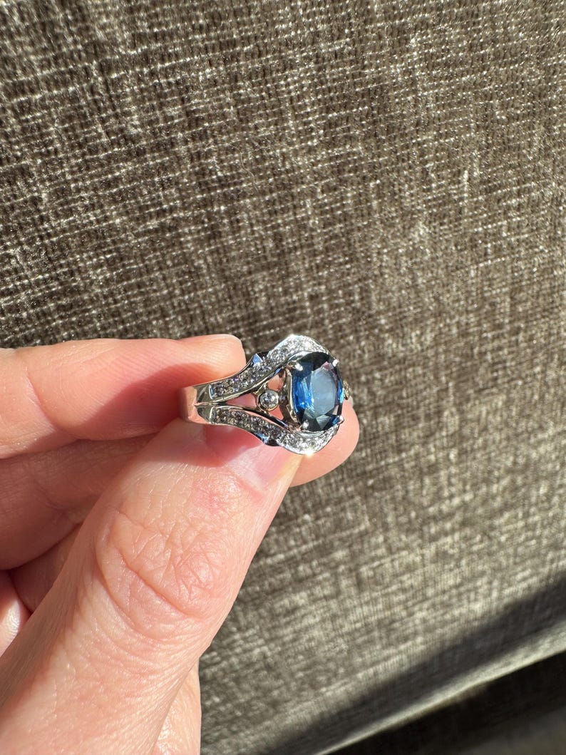 May include: A silver ring featuring a large, oval-cut, deep blue gemstone as the centerpiece. The ring is adorned with smaller, clear gemstones along the band and around the main stone, creating a sparkling effect. The ring is held up against a brown background.