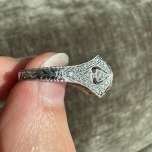 K&ouml;nnte beinhalten: Ein silberner Ring mit aufwendigem Filigran und kleinen, funkelnden Edelsteinen. Der Ring hat ein detailliertes Blumenmuster entlang des Bandes und ein zentrales Design mit mehreren Steinen. Der Ring wird hochgehalten, um seine verzierten Details zu zeigen.