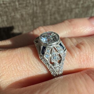 K&ouml;nnte beinhalten: Nahaufnahme eines silbernen Rings mit einem gro&szlig;en, klaren, oval geschliffenen Mittelstein. Der Ring weist aufwendige Filigranarbeiten und kleine Akzentsteine, darunter blaue Saphire, auf. Der Ring befindet sich an einem Finger und zeigt sein Design und seine Handwerkskunst.