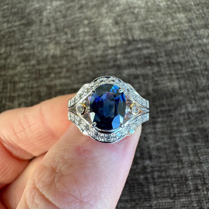 May include: A silver ring featuring a large, oval-cut, deep blue gemstone. The ring is accented with smaller, clear gemstones and gold-colored details, creating a luxurious and elegant appearance.