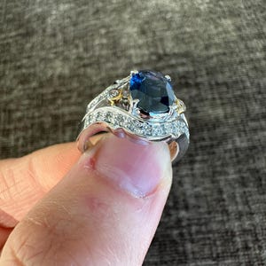 May include: A silver ring with a large, oval-cut, dark blue gemstone as the centerpiece. The ring is accented with smaller, clear gemstones and gold-colored details. The ring is held up by a finger.