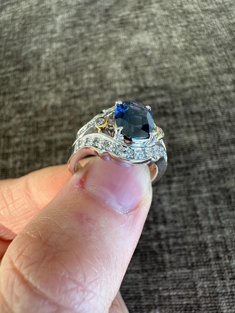May include: A silver ring with a large, oval-cut, dark blue gemstone as the centerpiece. The ring is accented with smaller, clear gemstones and gold-colored details. The ring is held up by a finger.