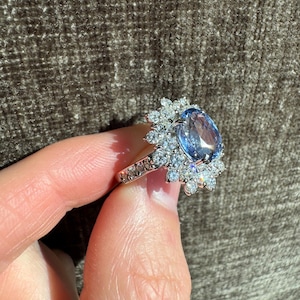 May include: A silver ring featuring a large, oval-cut, light blue gemstone surrounded by smaller, clear gemstones. The ring's band is also adorned with small, clear gemstones. The ring is held by a person's hand.