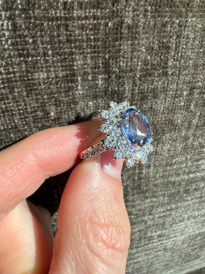 May include: A silver ring featuring a large, oval-cut, light blue gemstone surrounded by smaller, clear gemstones. The ring's band is also adorned with small, clear gemstones. The ring is held by a person's hand.