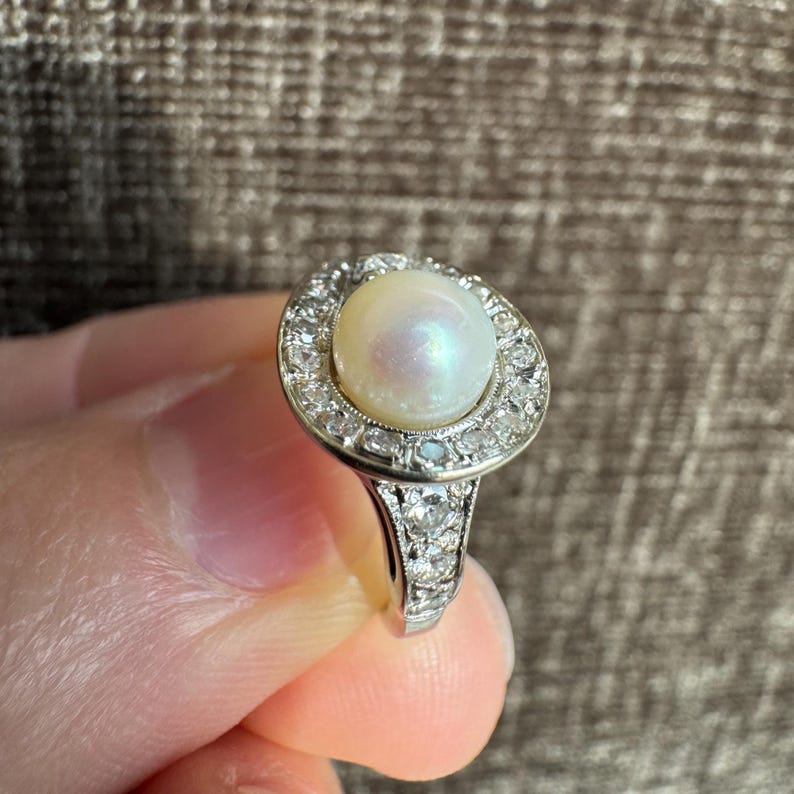 May include: A vintage-style ring featuring a large, luminous pearl at the center, surrounded by a halo of small, sparkling diamonds. The band is also adorned with diamonds, creating a classic and elegant design. The ring is held up by a hand.