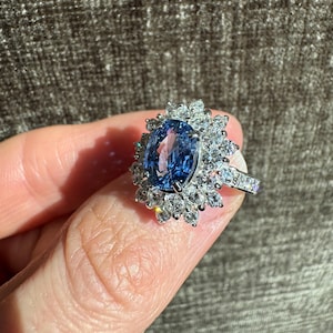 May include: A silver ring featuring a large, oval-cut blue gemstone surrounded by a halo of smaller, clear gemstones. The ring's band is also adorned with small, clear gemstones. The ring is held up by a finger.