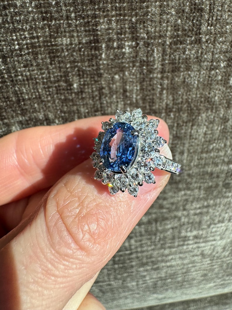 May include: A silver ring featuring a large, oval-cut blue gemstone surrounded by a halo of smaller, clear gemstones. The ring's band is also adorned with small, clear gemstones. The ring is held up by a finger.