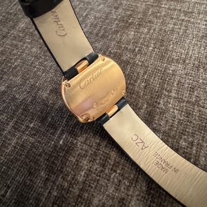 Puede incluir: Primer plano de un reloj Cartier dorado con una correa de cuero negro. La esfera del reloj es rectangular con bordes redondeados. La correa est&aacute; marcada "Made in France".