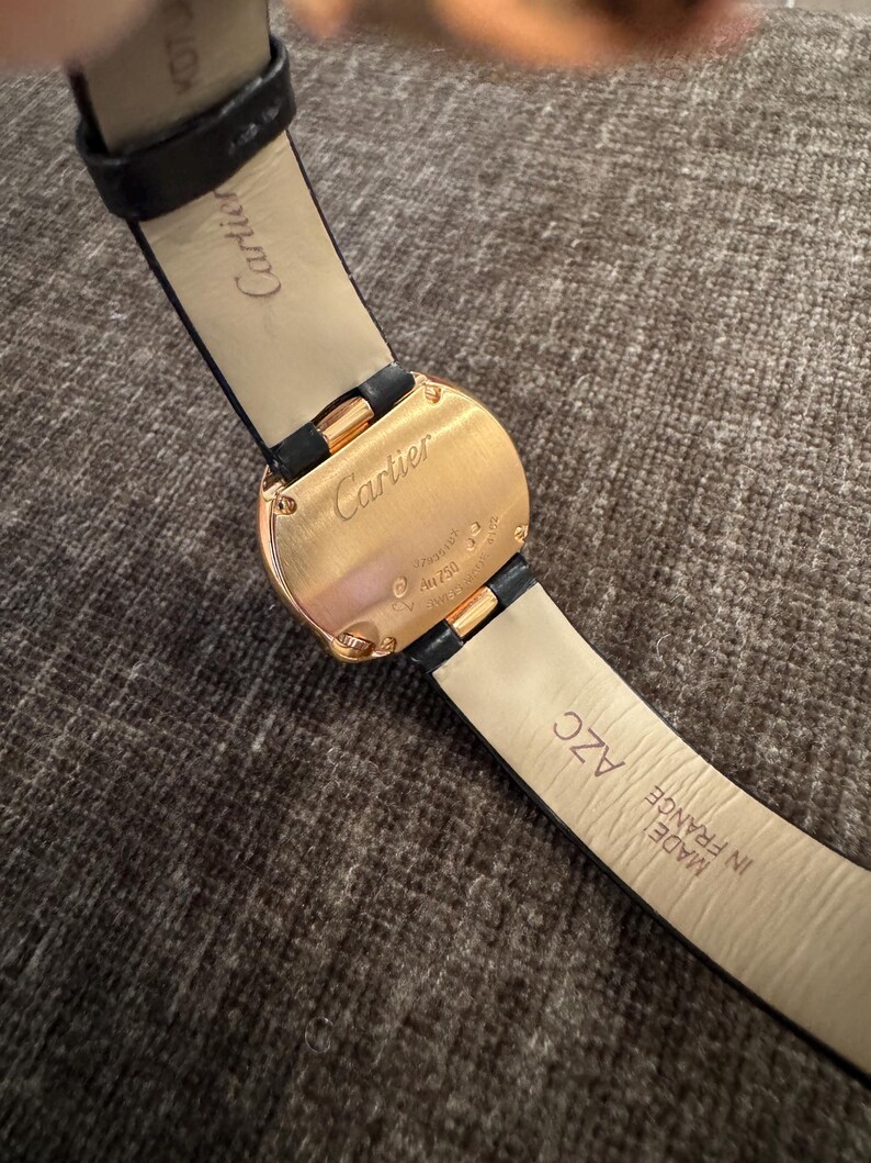 Puede incluir: Primer plano de un reloj Cartier dorado con una correa de cuero negro. La esfera del reloj es rectangular con bordes redondeados. La correa est&aacute; marcada "Made in France".