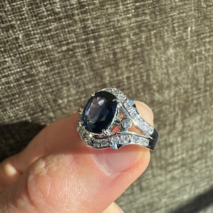 May include: A silver ring with a large, oval, dark blue gemstone as the centerpiece. The ring is encrusted with small, clear gemstones around the main stone and along the band. The ring has a detailed, ornate design, showcasing a luxurious aesthetic.