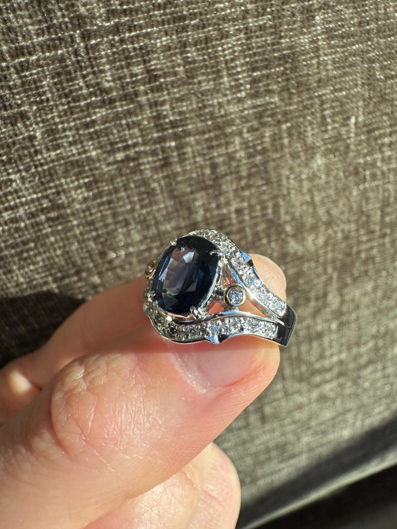 May include: A silver ring with a large, oval, dark blue gemstone as the centerpiece. The ring is encrusted with small, clear gemstones around the main stone and along the band. The ring has a detailed, ornate design, showcasing a luxurious aesthetic.