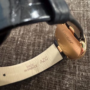 Puede incluir: Primer plano de un reloj Cartier dorado con correa de cuero negro. La esfera del reloj es visible y la correa est&aacute; marcada con "MADE IN FRANCE". El reloj tiene un dise&ntilde;o cl&aacute;sico y elegante.