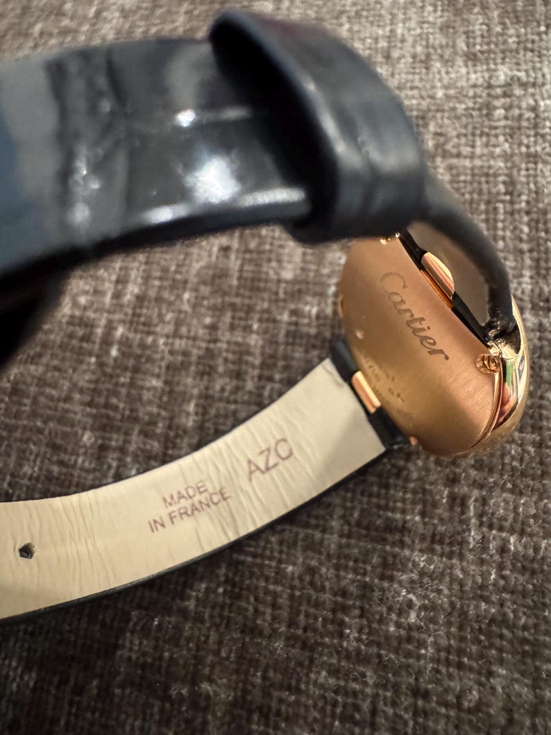 Puede incluir: Primer plano de un reloj Cartier dorado con correa de cuero negro. La esfera del reloj es visible y la correa est&aacute; marcada con "MADE IN FRANCE". El reloj tiene un dise&ntilde;o cl&aacute;sico y elegante.