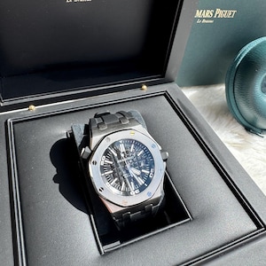 May include: A dark grey Audemars Piguet wristwatch with a textured black dial, luminous hands, and a metallic link bracelet, presented inside a black branded display box with the text Audemars Piguet Le Brassus printed in gold on the interior lid.