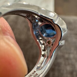 May include: A silver ring with a large, square-cut blue gemstone at the center. The ring is set with smaller, clear gemstones around the central stone. The band has a polished finish and appears to be made of a precious metal.