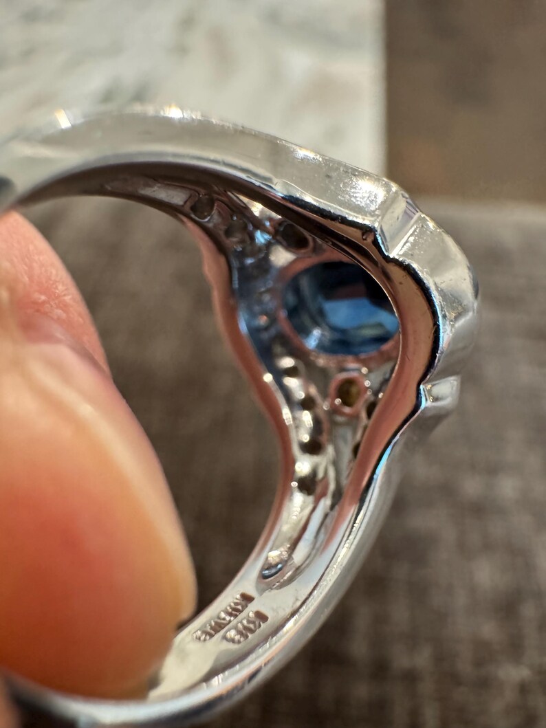 May include: A silver ring with a large, square-cut blue gemstone at the center. The ring is set with smaller, clear gemstones around the central stone. The band has a polished finish and appears to be made of a precious metal.