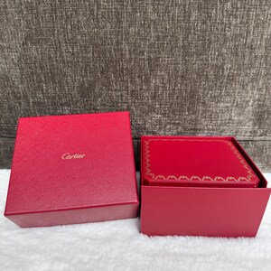 Puede incluir: Una caja roja de Cartier con el nombre de la marca en letras doradas. La caja est&aacute; abierta, revelando un interior rojo con un borde dorado. La caja est&aacute; sobre una superficie blanca.
