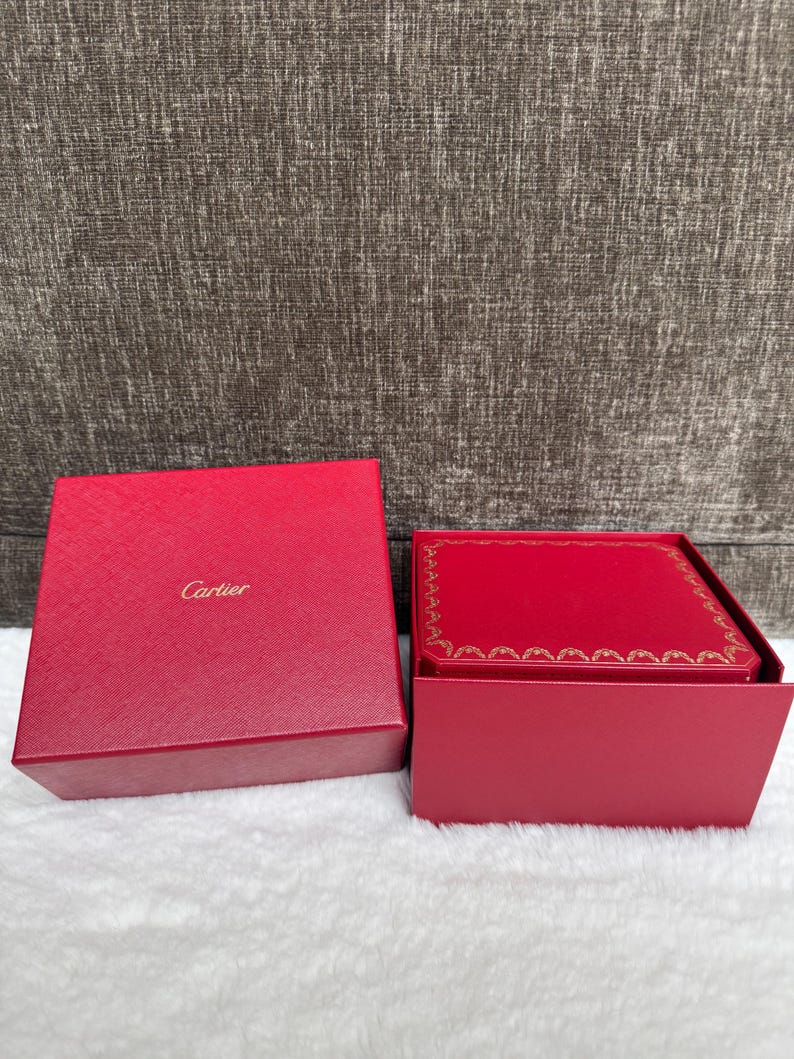 Puede incluir: Una caja roja de Cartier con el nombre de la marca en letras doradas. La caja est&aacute; abierta, revelando un interior rojo con un borde dorado. La caja est&aacute; sobre una superficie blanca.