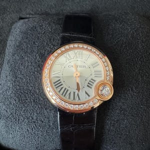 Puede incluir: Un reloj de lujo con una correa de cuero negro. La esfera es plateada con n&uacute;meros romanos y la marca "Cartier". El bisel es dorado e incrustado con diamantes. Un detalle con diamantes est&aacute; en el lateral.