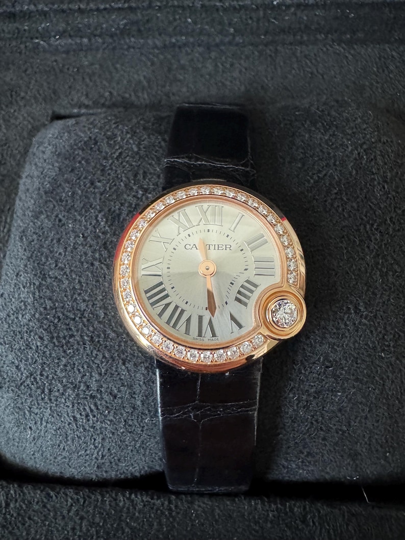 Puede incluir: Un reloj de lujo con una correa de cuero negro. La esfera es plateada con n&uacute;meros romanos y la marca "Cartier". El bisel es dorado e incrustado con diamantes. Un detalle con diamantes est&aacute; en el lateral.