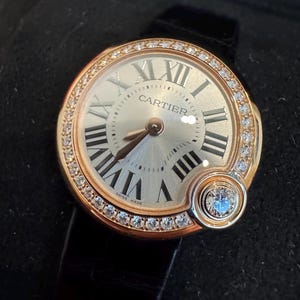 Puede incluir: Un reloj de pulsera dorado con esfera redonda, n&uacute;meros romanos y la marca "CARTIER". El bisel est&aacute; engastado con peque&ntilde;os diamantes, y un diamante m&aacute;s grande adorna el lateral. El reloj tiene una correa de cuero negro.