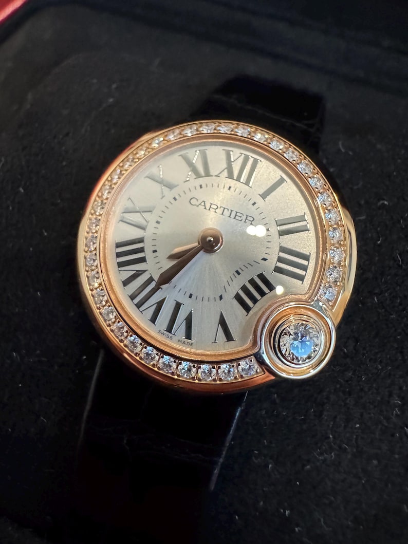 Puede incluir: Un reloj de pulsera dorado con esfera redonda, n&uacute;meros romanos y la marca "CARTIER". El bisel est&aacute; engastado con peque&ntilde;os diamantes, y un diamante m&aacute;s grande adorna el lateral. El reloj tiene una correa de cuero negro.