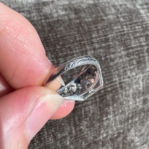K&ouml;nnte beinhalten: Nahaufnahme eines verzierten Silberrings mit aufwendigen floralen Gravuren. Der Ring hat eine zentrale Anordnung kleiner, funkelnder Edelsteine, m&ouml;glicherweise Diamanten, und kleinere blaue Steine. Der Ring wird zwischen Fingern gehalten, was sein detailliertes Design zeigt.