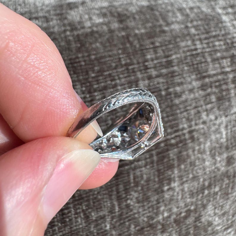 K&ouml;nnte beinhalten: Nahaufnahme eines verzierten Silberrings mit aufwendigen floralen Gravuren. Der Ring hat eine zentrale Anordnung kleiner, funkelnder Edelsteine, m&ouml;glicherweise Diamanten, und kleinere blaue Steine. Der Ring wird zwischen Fingern gehalten, was sein detailliertes Design zeigt.