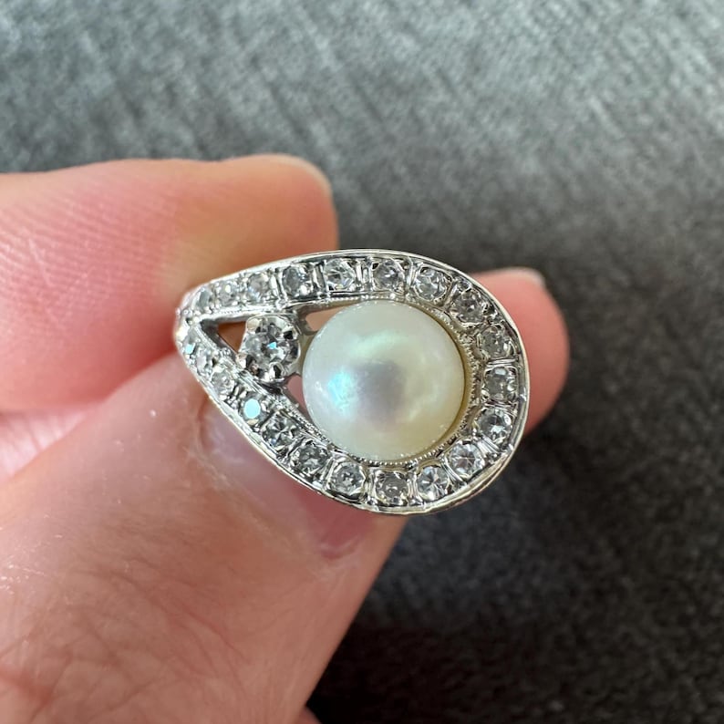 May include: A vintage-style ring featuring a large, lustrous pearl at the center, surrounded by a halo of small, sparkling diamonds. The ring has a unique, teardrop-shaped design, with additional diamond accents on the side. The metal appears to be silver or white gold.