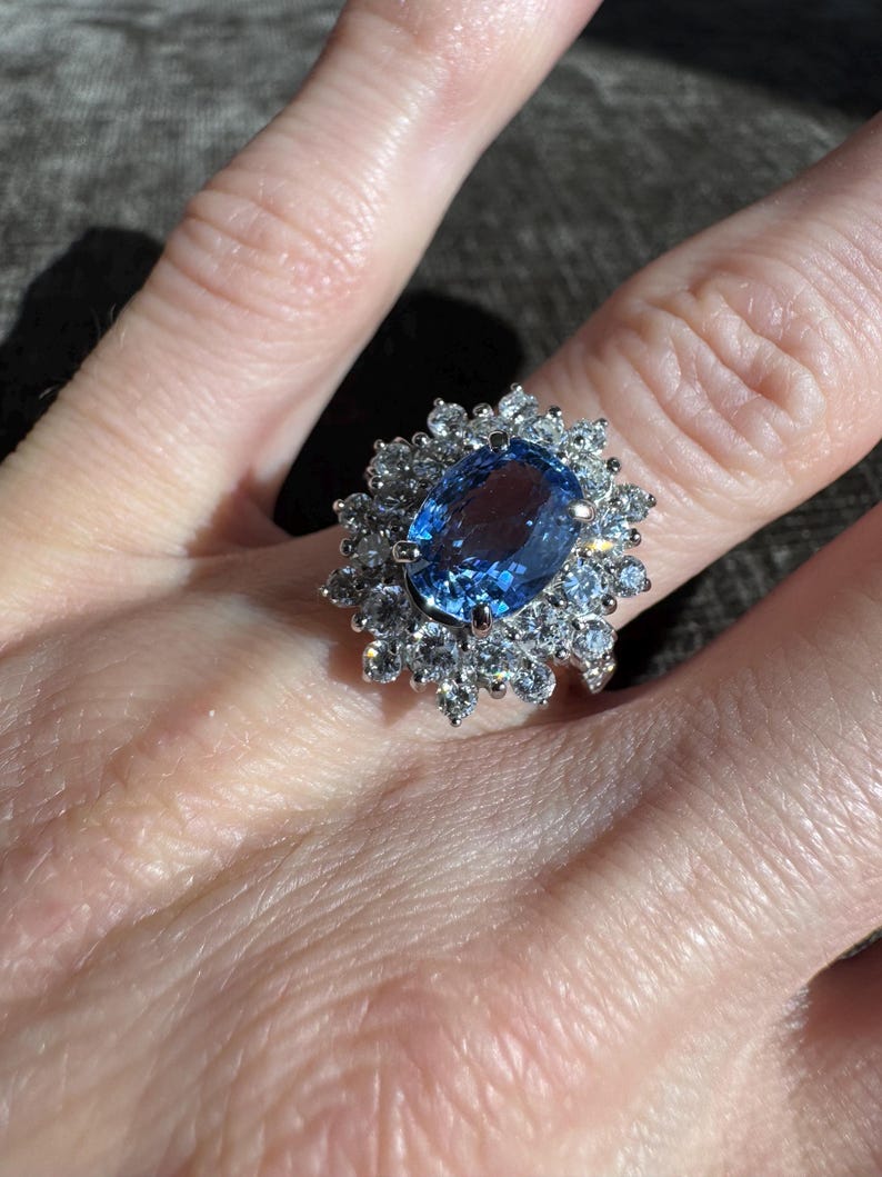 May include: A silver ring featuring a large, oval-cut blue gemstone surrounded by smaller, clear gemstones. The ring's design is a cluster of stones, creating a sparkling, eye-catching effect. The ring is shown on a person's finger.