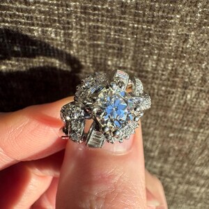 Może przedstawiać: Olśniewający pierścionek w stylu vintage z dużym, okrągłym diamentem w centrum. Diament otoczony jest mniejszymi, błyszczącymi kamieniami i skomplikowaną metaloplastyką. Pierścionek trzymany jest przez palec.