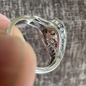 May include: Silver ring with a heart-shaped design, featuring a central yellow stone and small clear stones along the edges. The ring is held by a person's finger, showcasing the intricate details of the setting.